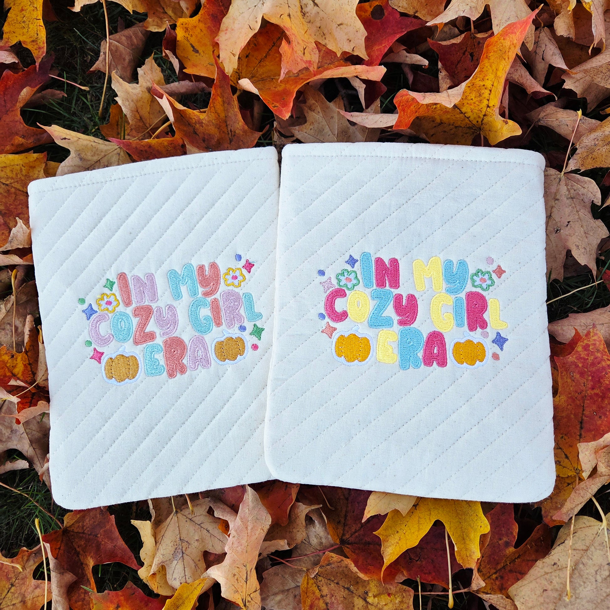 Two quilted Kindle sleeves embroidered with “In My Cozy Girl Era” surrounded by fall leaves in autumn colors.
